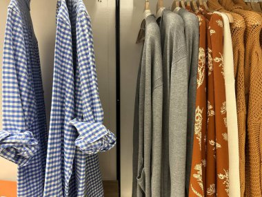 Mavi kareli gömlekler, gri kazaklar, turuncu çiçek desenli bluzlar ve turuncu örgü giysiler de dahil olmak üzere askıda asılı giysiler.