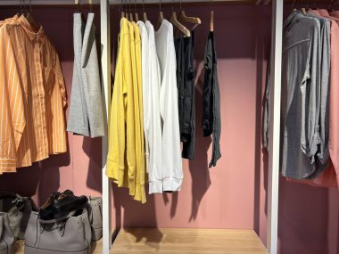 Bir dolap, altında gri çantalar ve siyah ayakkabılar bulunan çeşitli düzgün asılmış giysiler barındırır.