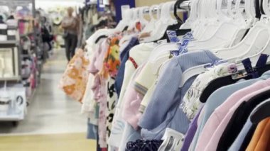 Bir perakende mağazasında askılarda renkli çocuk kıyafetleri, arka planda çeşitli ev eşyaları görünür