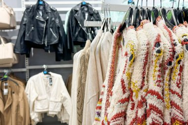 Bir giyim mağazasında askılarda asılı duran çeşitli ceket ve kazaklar ve arka planda el çantaları sergilenmektedir.