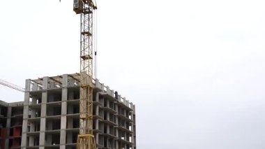 Uzun sarı bir vinç bulutlu bir gökyüzünün altında kısmen inşa edilmiş çok katlı beton bir binanın yanında duruyor.
