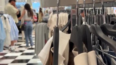 Askıların üzerindeki nötr renkli giysiler odak noktasında sergilenirken, insanların yer aldığı canlı bir moda mağazası sahnesi