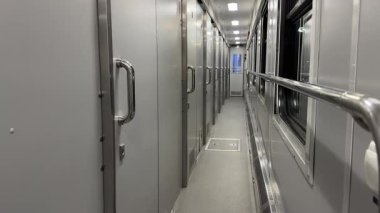 Modern bir tren vagonunda uzun dar bir koridor metalik kapı ve pencereleri gösteriyor.