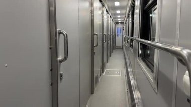 Modern bir tren vagonunda uzun dar bir koridor metalik kapı ve pencereleri gösteriyor.