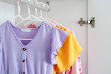 Renkli çocuk kıyafetleri, fırfırlı kolları olan leylak bir elbise, dolaptaki beyaz askılara asılı.