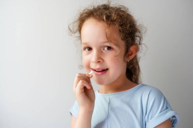 Açık mavi tişörtlü, gülümseyen bir kız çocuğu ağzının yanında beyaz bir tablet tutuyor ve almaya hazır görünüyor.