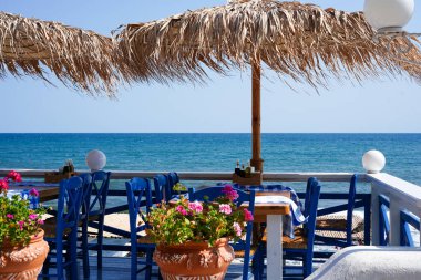 Mavi sandalyeli deniz kenarı restoranı, kareli masa örtüleri ve sakin okyanusa bakan canlı çiçekler.