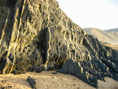 Muazzam bir kaya oluşumu kumlu bir plajda dimdik duruyor, engin bir gökyüzünün altında yükselen dağlar var.