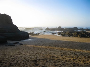 Uçsuz bucaksız okyanusun arka planında kayalarla süslenmiş kumlu bir sahil, doğal ve okyanus manzaralarının güzelliğini gözler önüne seriyor.