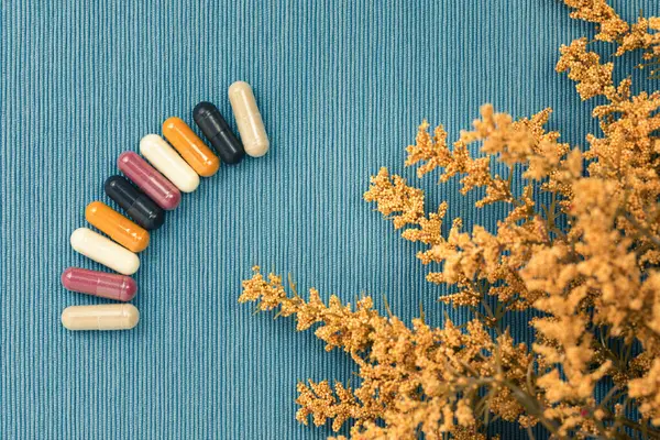 Top view of a variety of vitamin and mineral supplements in capsules. Multicolored pills in orange, red, white, black on a blue textured background. Decorative dried plant in golden brown.