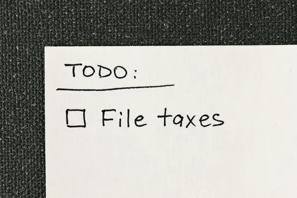 Handwritten to-do list with \'File taxes\' reminder and a checkbox written on a white note against a gray textured backdrop, flat lay top-down view