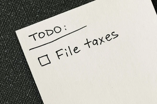 Hand written to-do list with 'File taxes' reminder and a checkbox written on a white note against a gray textured backdrop, flat lay top down view