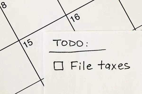 Hand written to-do list with 'File taxes' reminder written on a sticky note over a calendar, with income tax return filing deadline of April 15