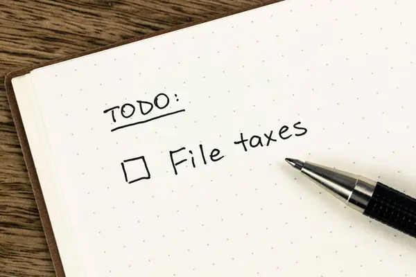 To-do list with \'File taxes\' checkbox reminder handwritten on an open dot grid notebook with a pen on a textured wooden desk, flat lay, top-down view