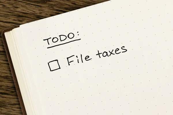 To-do list with \'File taxes\' checkbox reminder handwritten on an open dot grid notebook on a textured wooden desk, flat lay, top-down view