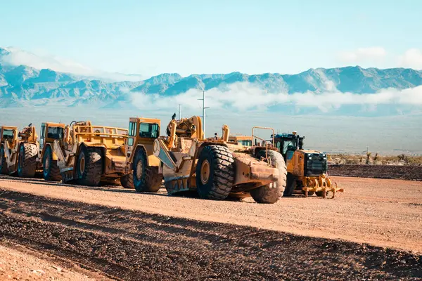 Toprak taşıyıcılardan oluşan bir filo, yeni konut geliştirme çalışmaları için çorak çöl arazisi hazırlıyor. İnşaat ekipmanları engebeli araziyi yerleşim alanı olarak değiştirirken, arka planda dağlar beliriyor..