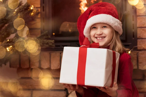 Christmas Child Girl in Santa Hat with Big Gift Box or Red Present near Christmas Tree with Lights and Fireplace with Copy Space. Merry Christmas for Children