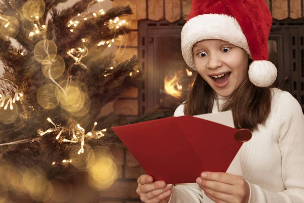 Surprised Christmas Girl Teenager Received Christmas Gift Envelope with Christmas Certificate near Tree and Fireplace.  Wow Face of Teen in Christmas Eve