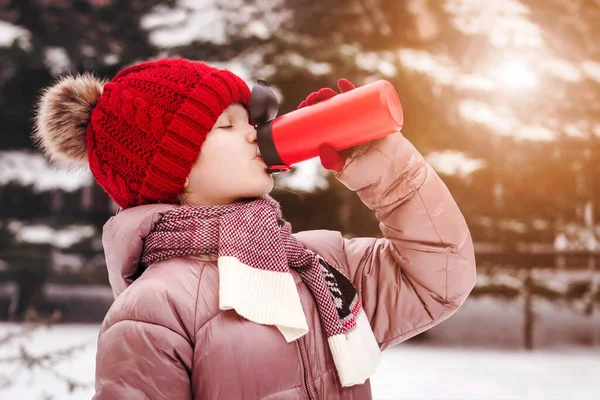 Thermos for Children. Child Girl Drinking Hot Drink from Thermos Mug in Cold Snow Winter Park.