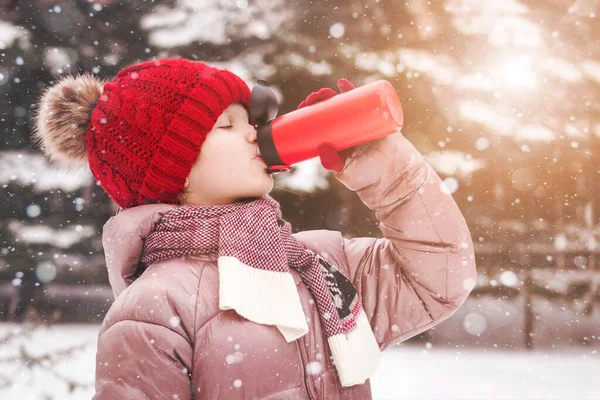 Thermos or Thermal mug for Traveler Teenager or Children in Cold Winter. Girl Teen Drinking Hot Tea or Coffee from Red Thermos Mug in Snow Windows Outside.
