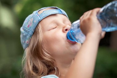 Drinking Water from Bottle. Thirsty Child Little Girl Baby Drink Water on Green Nature Background Outside.