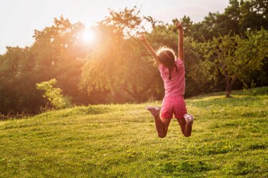 Yaz Tatili 'nde Doğa Arkaplanı' nda iyi eğlenceler. Küçük kız zıplıyor Yeşil Çimenlikte Elleri Gün Batımında Keyifli bir hayat sürüyor.