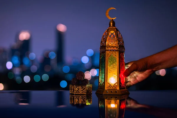 Lantern holding by hand and small plate of dates fruit on table with night sky and city bokeh light background for the Muslim feast of the holy month of Ramadan Kareem.