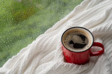 Bir fincan sıcak kahve beyaz battaniye ve pencereye yağmur damlası koyar. Evde kal ve konsepti rahatlat..