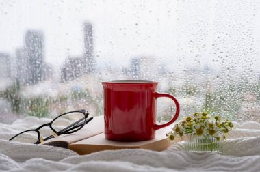 A red mug of coffee and spectacles put on book when rain drop on window. Stay home and relaxing concept.	