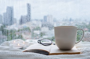 A coffee mug and spectacles put on book with candle on blanket when rain drop on window. Stay home and relaxing concept.	