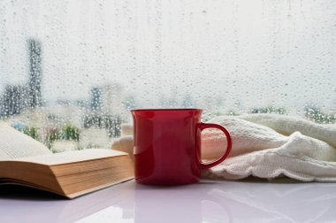 Sabahları üzerinde kitap ve battaniye olan kırmızı bir fincan kahve ve penceresinde yağmur damlası. Evde kal ve konsepti rahatlat..