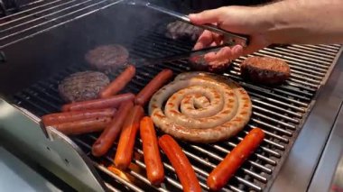Bir adam açık hava gaz ızgarasında sosisli, hamburger ve sarmal sosis pişiriyor. Hafta sonu pikniğinde maşa kullanarak sosisleri çeviriyor..