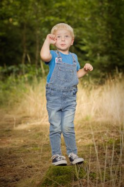 Kot giymiş küçük bir çocuk ormanda kütük ve patika üzerinde poz veriyor.