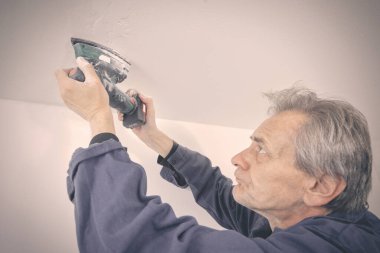 Yaşlı adam boş bir apartmanın tavanını tamir edip zımparalıyor.
