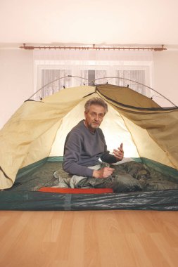 Yaşı büyük bir adam dışarıda kamp yapmak yerine apartman odasında çadır kuruyor.