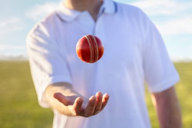 Cricket player bowler throwing up and catching red leather cricket ball ready to bowl at cricket pitch clipart