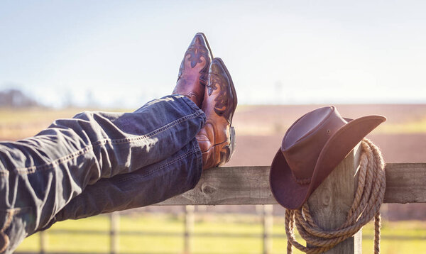 Cowboy boots and hat with feet up on stables fence at ranch resting with legs crossed, country music festival live concert or line dancing concept