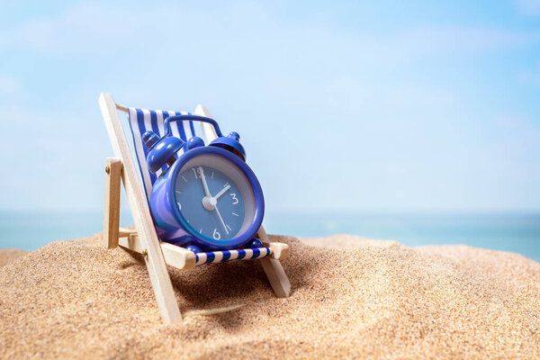 Time to relax, alarm clock relaxing on deck chair on sand beach with sea and sky background