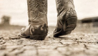 Çiftlikteki kurak arazide arka planda ahırlarla birlikte yürüyen kovboy çizmeleri.