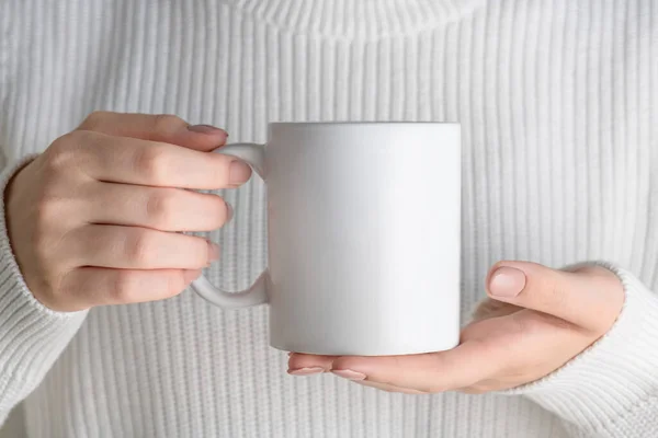 Female hand holding white mug mockup with blank copy space for your advertising text message or promotional content. Girl in blue shirt holding white porcelain coffee mug mock up