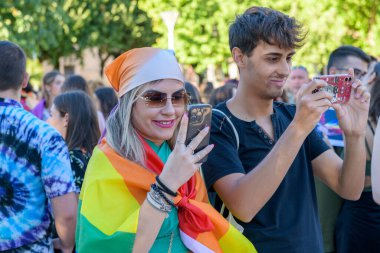 Manduria, İtalya - 13 Temmuz 2022. Gökkuşağı bayrağı takan bir kız ve bir adam LGBT gay yürüyüşü sırasında akıllı telefonuyla fotoğraf çekiyorlar. Sokakta LGBTQ onur günü kutlamaları