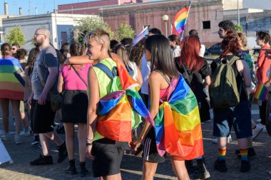 Manduria, İtalya - 13 Temmuz 2022. Genç, gülümseyen lezbiyen çift, LGBTQ eşcinsellik yürüyüşü sırasında sırtlarında gökkuşağı bayrağıyla Güney İtalya 'da seçici bir odak noktasında yürüyor.