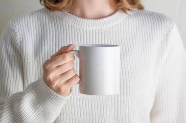 Female hands holding white mug mockup with blank copy space for your advertising text message or promotional content. Girl in white sweater holding white porcelain coffee mug mock up, close up