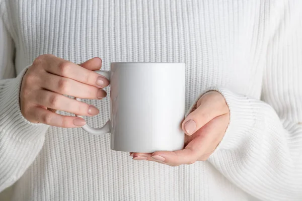 Female hands holding white mug mockup with blank copy space for your advertising text message or promotional content. Girl in white sweater holding white porcelain coffee mug mock up, close up