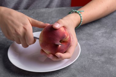 Otantik kadın elleri, gri masa arkaplanındaki beyaz tabakta taze bir şeftaliyi kesiyor, yakın plan. Sağlıklı yaz atıştırması, yemek yaşam tarzı.