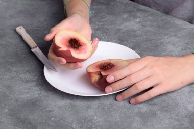 Gri masa arkaplanındaki beyaz tabakta taze şeftalinin yarısını tutan otantik kadın elleri, yakın plan. Sağlıklı yaz atıştırması, yemek yaşam tarzı.