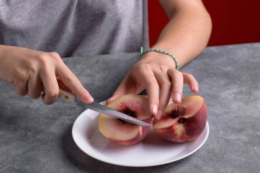 Otantik kadın elleri, gri masa arkaplanındaki beyaz tabakta taze, olgun bir şeftaliyi kesiyor, yakın plan. Sağlıklı yaz atıştırması, yemek yaşam tarzı.