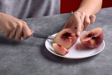 Otantik kadın elleri, gri masa arkaplanındaki beyaz tabakta taze, olgun bir şeftaliyi kesiyor, yakın plan. Sağlıklı yaz atıştırması, yemek yaşam tarzı.