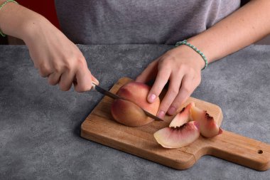 Otantik kadın elleri, gri masa arkaplanındaki tahta kesme tahtasıyla taze olgun bir şeftaliyi kesiyor. Yaklaş. Sağlıklı yaz atıştırması, yemek yaşam tarzı.