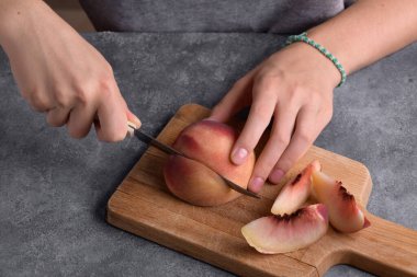 Otantik kadın elleri, gri masa arkaplanındaki tahta kesme tahtasıyla taze olgun bir şeftaliyi kesiyor. Yaklaş. Sağlıklı yaz atıştırması, yemek yaşam tarzı.